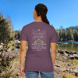 Purple Christian t-shirt with Isaiah 9:6 scripture, stars and barn design, worn by a person standing by a lake with pine trees under a blue sky.