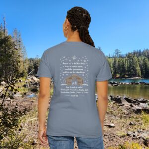 Blue Christian t-shirt with Isaiah 9:6 scripture, stars and barn design, worn by a person standing by a lake with pine trees under a blue sky.