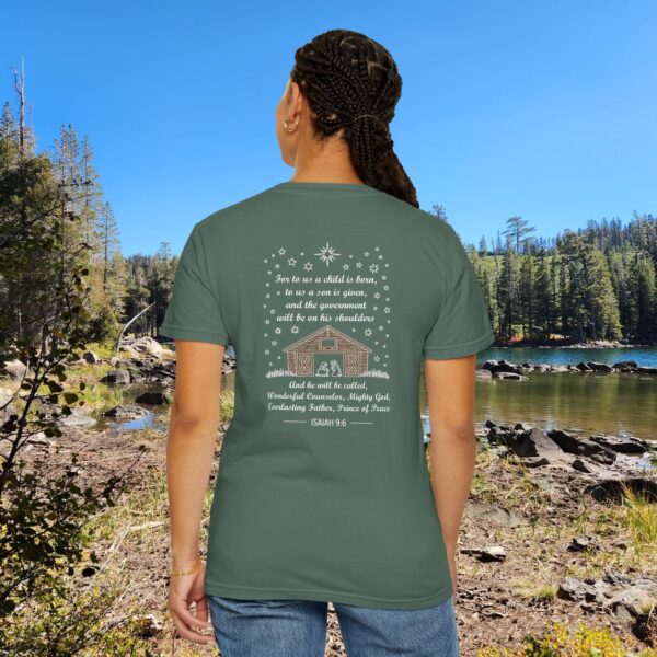 Green Christian t-shirt with Isaiah 9:6 scripture, stars and barn design, worn by a person standing by a lake with pine trees under a blue sky.