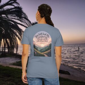 Blue Christian t-shirt with Jeremiah 29:13 scripture, mountain valley, and sunset graphic, worn by a person in a serene beach setting.