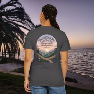 Gray Christian t-shirt with Jeremiah 29:13 scripture, mountain valley, and sunset graphic, worn by a person in a serene beach setting.