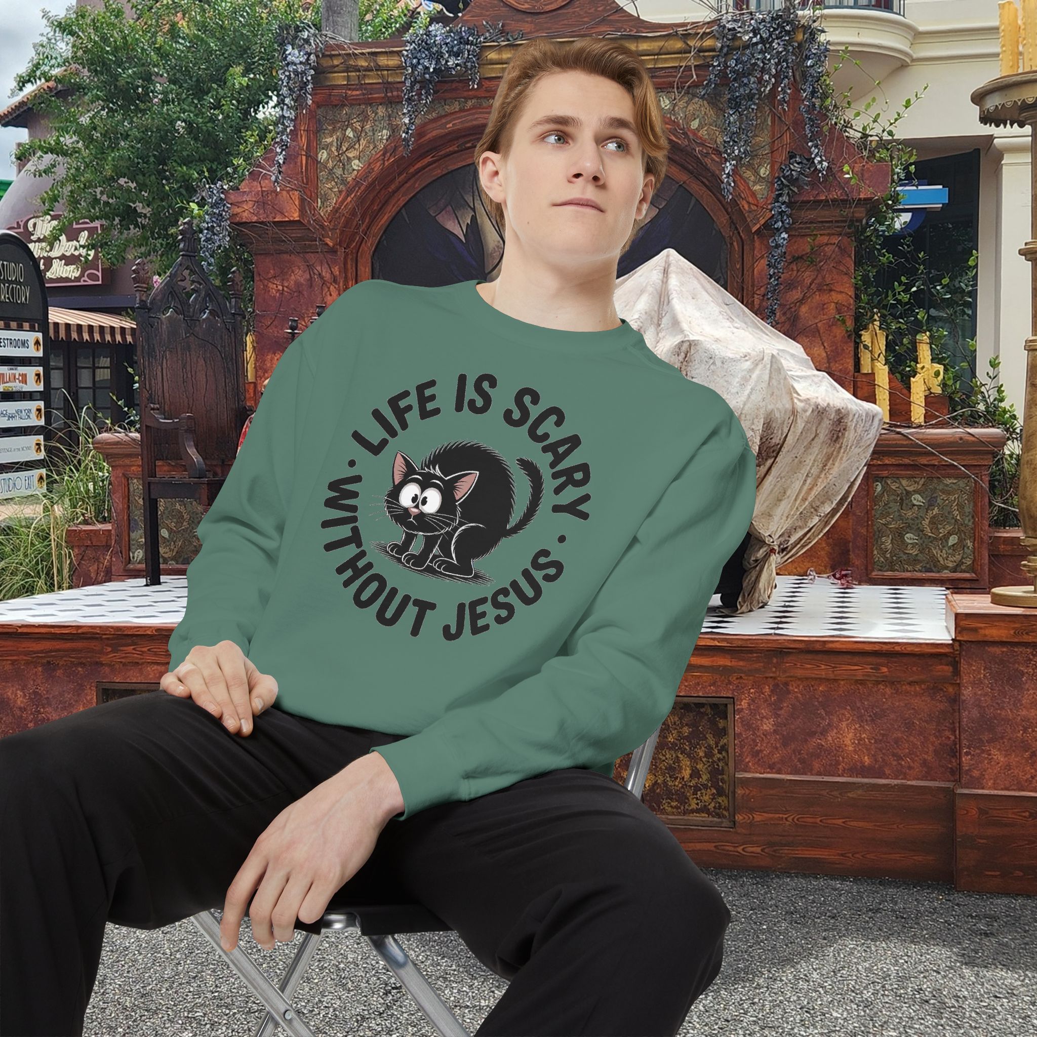 Model on a Halloween set wearing a green Life is Scary Without Jesus Halloween Sweatshirt, design showing a cute frightened black cat.