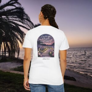 White Christian t-shirt with John 8:12 scripture, lit stairway path with flowers and sunset design, worn by a person standing on a sunset beach.