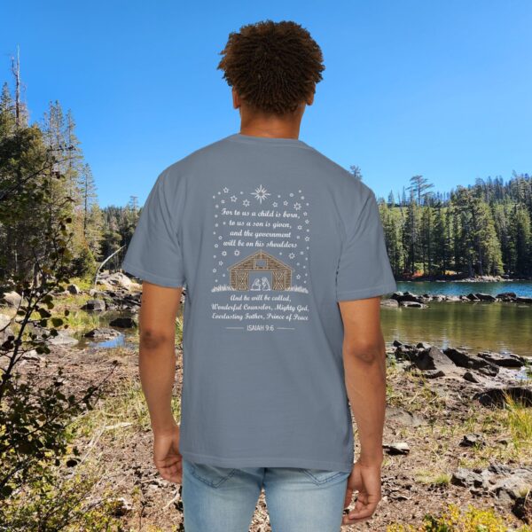Blue Christian t-shirt with Isaiah 9:6 scripture, stars and barn design, worn by a person standing by a lake with pine trees under a blue sky.
