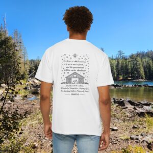 White Christian t-shirt with Isaiah 9:6 scripture, stars and barn design, worn by a person standing by a lake with pine trees under a blue sky.