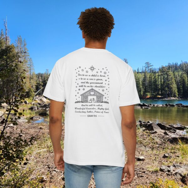 White Christian t-shirt with Isaiah 9:6 scripture, stars and barn design, worn by a person standing by a lake with pine trees under a blue sky.