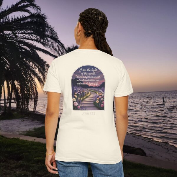 Ivory Christian t-shirt with John 8:12 scripture, lit stairway path with flowers and sunset design, worn by a person standing on a sunset beach.