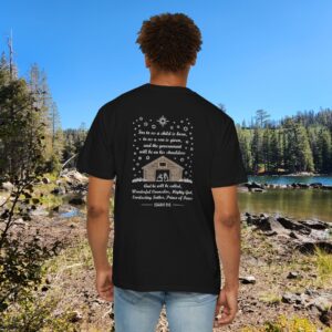 Black Christian t-shirt with Isaiah 9:6 scripture, stars and barn design, worn by a person standing by a lake with pine trees under a blue sky.