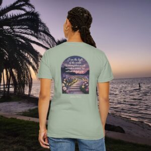 Green Christian t-shirt with John 8:12 scripture, lit stairway path with flowers and sunset design, worn by a person standing on a sunset beach.