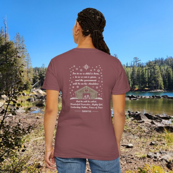 Maroon Christian t-shirt with Isaiah 9:6 scripture, stars and barn design, worn by a person standing by a lake with pine trees under a blue sky.
