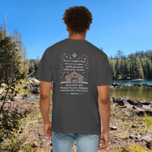 Gray Christian t-shirt with Isaiah 9:6 scripture, stars and barn design, worn by a person standing by a lake with pine trees under a blue sky.