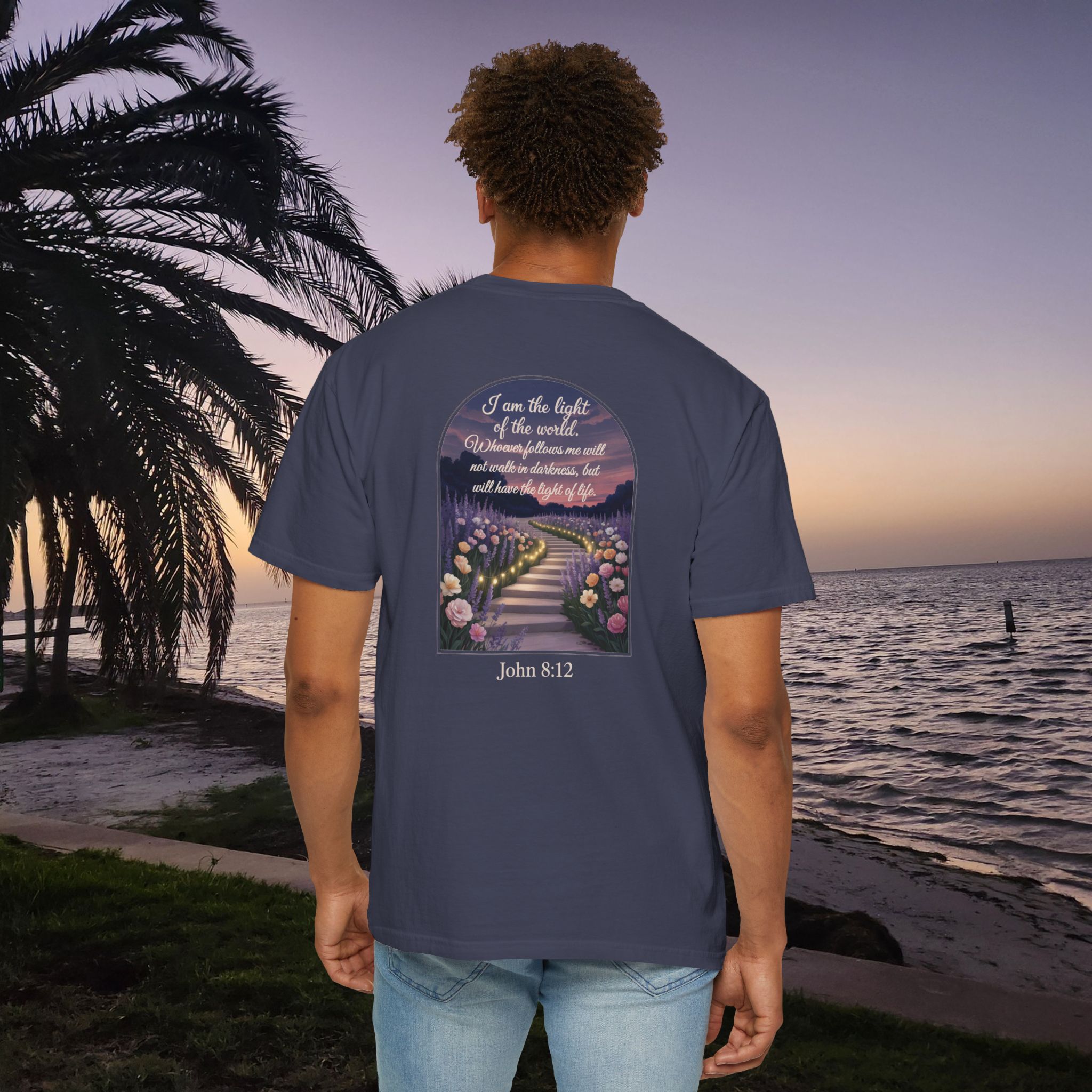 Blue Christian t-shirt with John 8:12 scripture, lit stairway path with flowers and sunset design, worn by a person standing on a sunset beach.