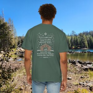 Green Christian t-shirt with Isaiah 9:6 scripture, stars and barn design, worn by a person standing by a lake with pine trees under a blue sky.