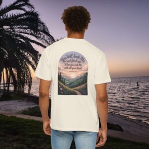 Ivory Christian t-shirt with Jeremiah 29:13 scripture, mountain valley, and sunset graphic, worn by a person in a serene beach setting.
