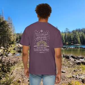 Purple Christian t-shirt with Isaiah 9:6 scripture, stars and barn design, worn by a person standing by a lake with pine trees under a blue sky.