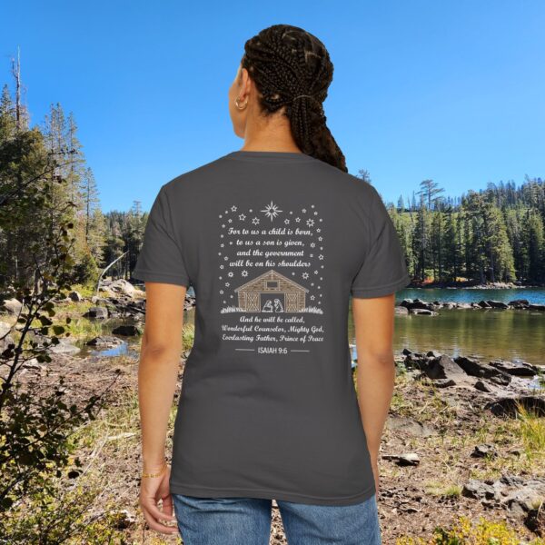 Gray Christian t-shirt with Isaiah 9:6 scripture, stars and barn design, worn by a person standing by a lake with pine trees under a blue sky.