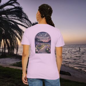 Pink Christian t-shirt with John 8:12 scripture, lit stairway path with flowers and sunset design, worn by a person standing on a sunset beach.