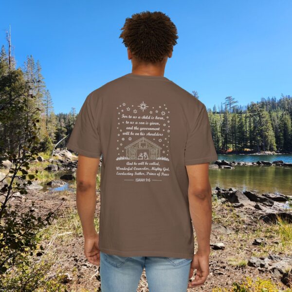 Brown Christian t-shirt with Isaiah 9:6 scripture, stars and barn design, worn by a person standing by a lake with pine trees under a blue sky.