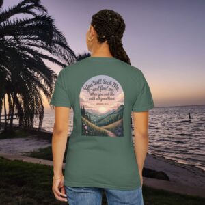 Green Christian t-shirt with Jeremiah 29:13 scripture, mountain valley, and sunset graphic, worn by a person in a serene beach setting.