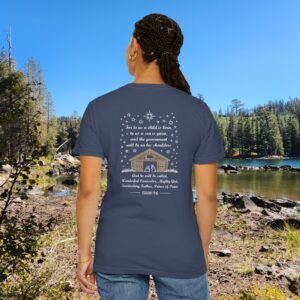 Blue Christian t-shirt with Isaiah 9:6 scripture, stars and barn design, worn by a person standing by a lake with pine trees under a blue sky.