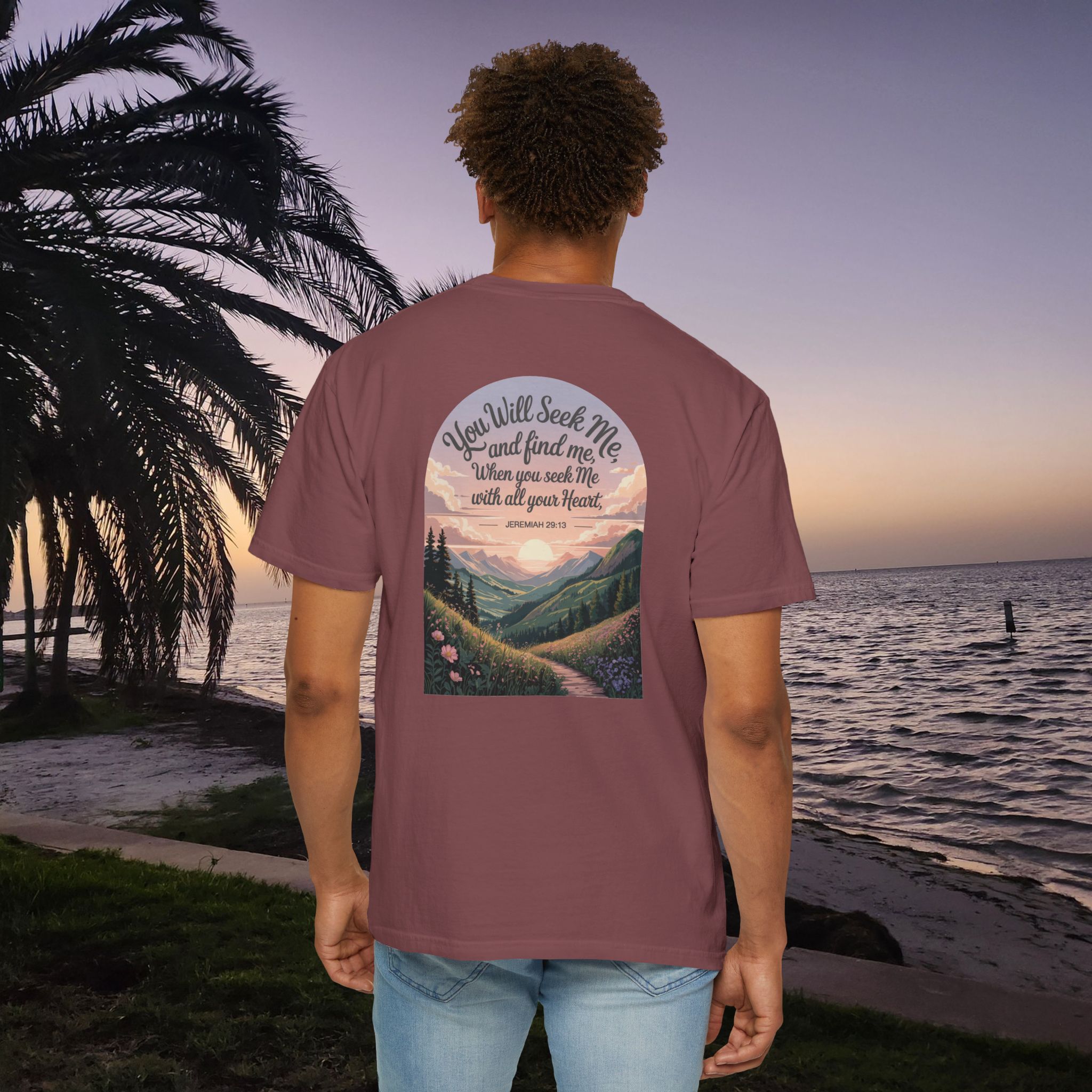 Red Christian t-shirt with Jeremiah 29:13 scripture, mountain valley, and sunset graphic, worn by a person in a serene beach setting.