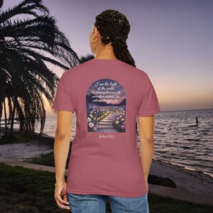 Pink Christian t-shirt with John 8:12 scripture, lit stairway path with flowers and sunset design, worn by a person standing on a sunset beach.