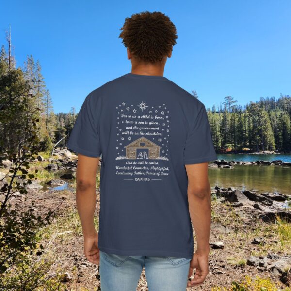 Blue Christian t-shirt with Isaiah 9:6 scripture, stars and barn design, worn by a person standing by a lake with pine trees under a blue sky.
