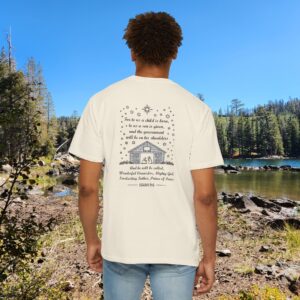 Ivory Christian t-shirt with Isaiah 9:6 scripture, stars and barn design, worn by a person standing by a lake with pine trees under a blue sky.