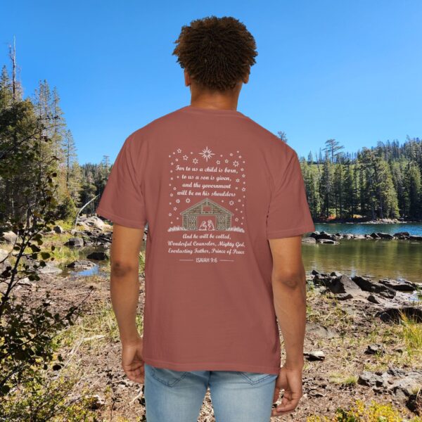 Maroon Christian t-shirt with Isaiah 9:6 scripture, stars and barn design, worn by a person standing by a lake with pine trees under a blue sky.