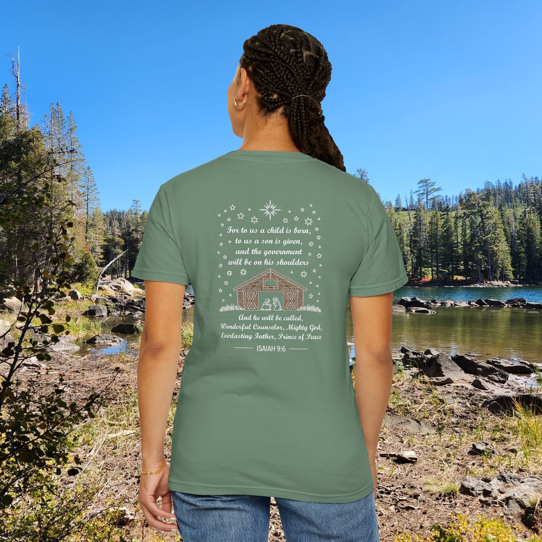 Green Christian t-shirt with Isaiah 9:6 scripture, stars and barn design, worn by a person standing by a lake with pine trees under a blue sky.