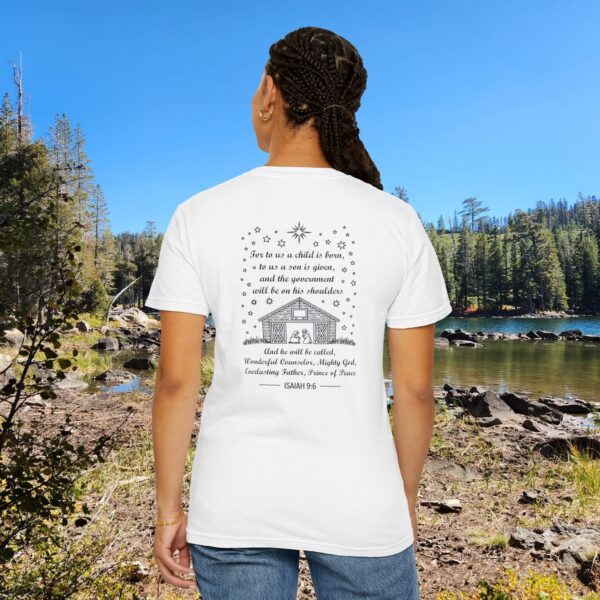 White Christian t-shirt with Isaiah 9:6 scripture, stars and barn design, worn by a person standing by a lake with pine trees under a blue sky.