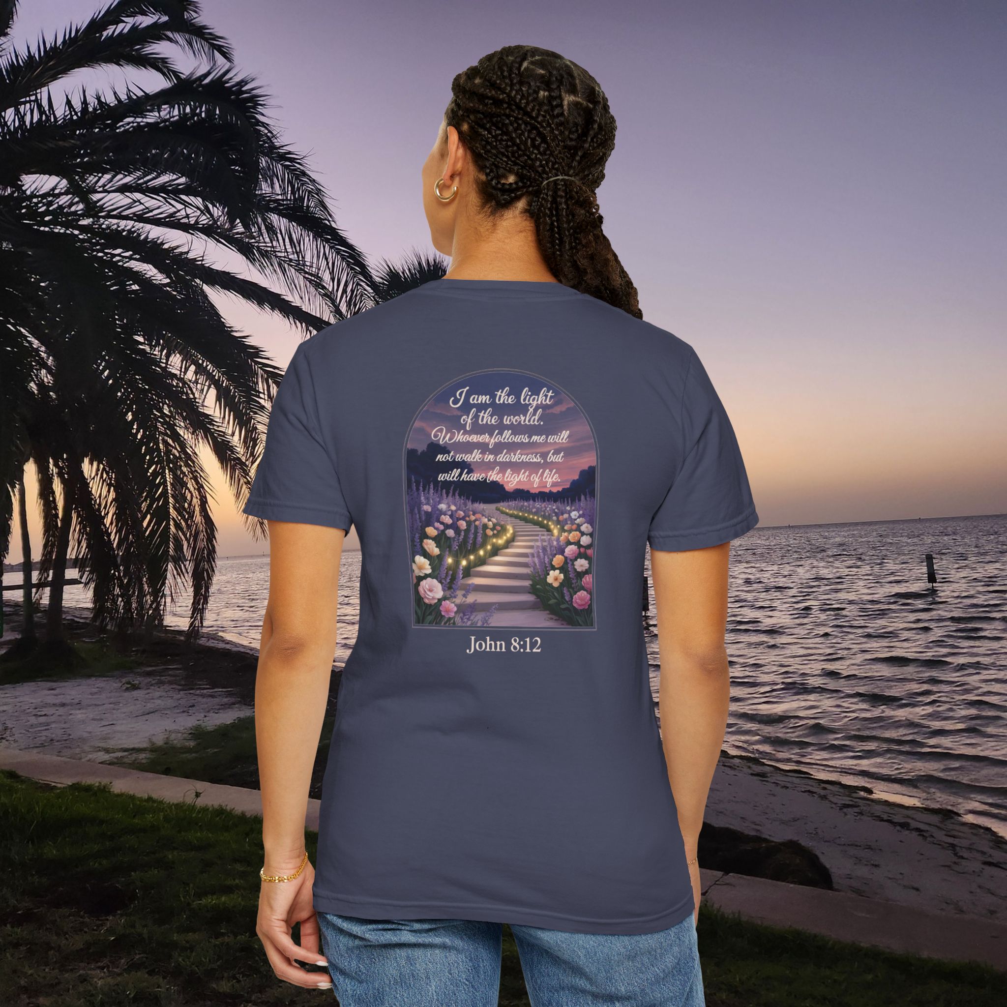 Blue Christian t-shirt with John 8:12 scripture, lit stairway path with flowers and sunset design, worn by a person standing on a sunset beach.