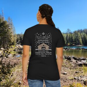 Black Christian t-shirt with Isaiah 9:6 scripture, stars and barn design, worn by a person standing by a lake with pine trees under a blue sky.