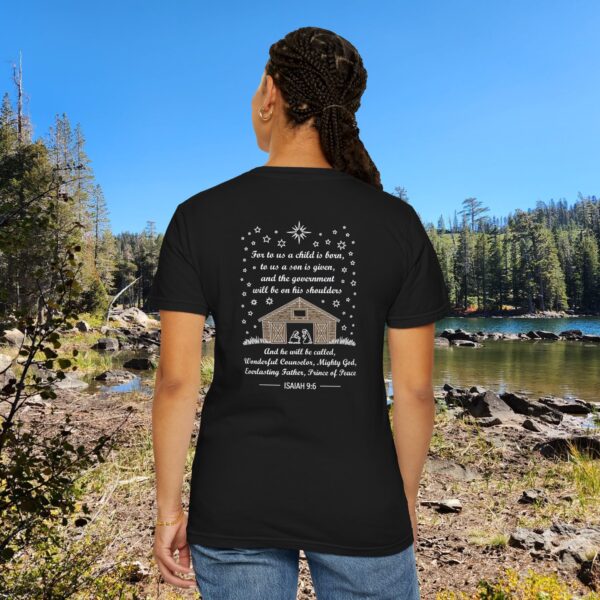 Black Christian t-shirt with Isaiah 9:6 scripture, stars and barn design, worn by a person standing by a lake with pine trees under a blue sky.