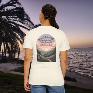 Ivory Christian t-shirt with Jeremiah 29:13 scripture, mountain valley, and sunset graphic, worn by a person in a serene beach setting.