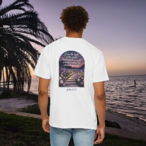White Christian t-shirt with John 8:12 scripture, lit stairway path with flowers and sunset design, worn by a person standing on a sunset beach.