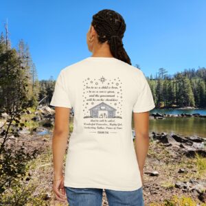 Ivory Christian t-shirt with Isaiah 9:6 scripture, stars and barn design, worn by a person standing by a lake with pine trees under a blue sky.