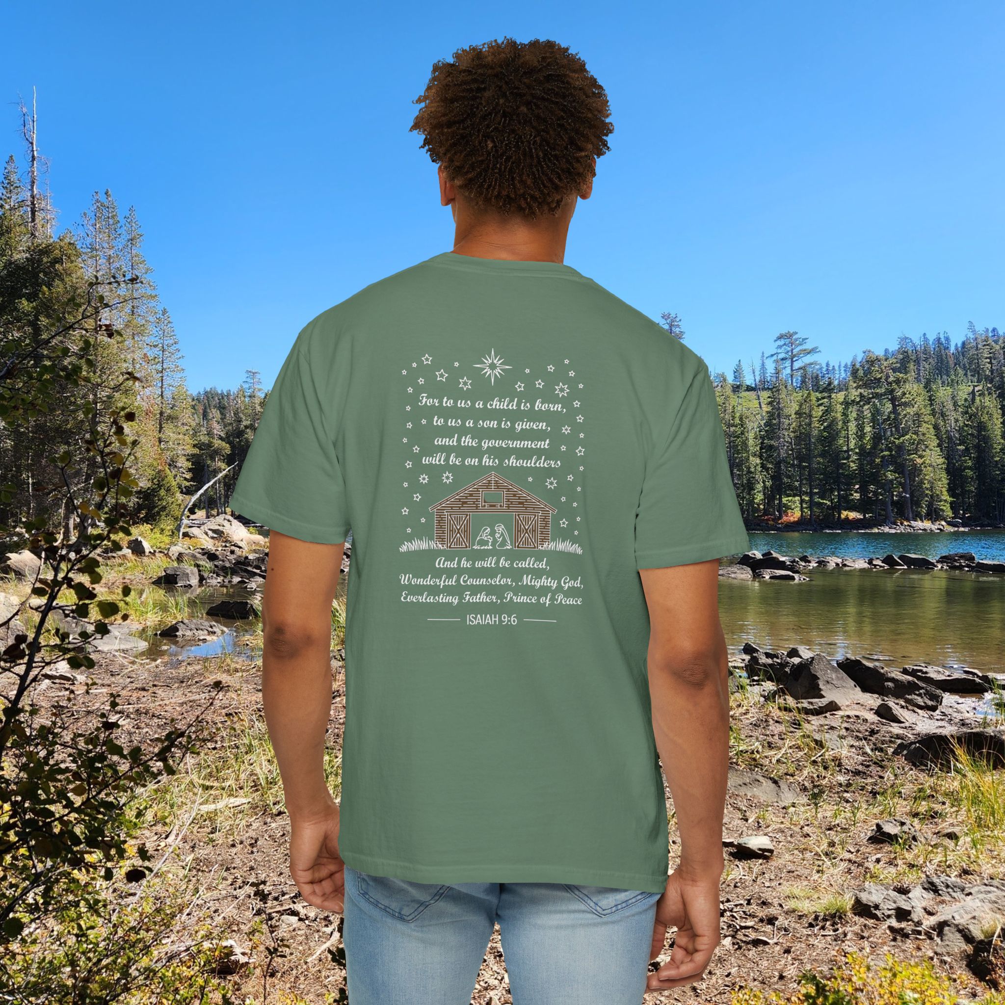 Green Christian t-shirt with Isaiah 9:6 scripture, stars and barn design, worn by a person standing by a lake with pine trees under a blue sky.