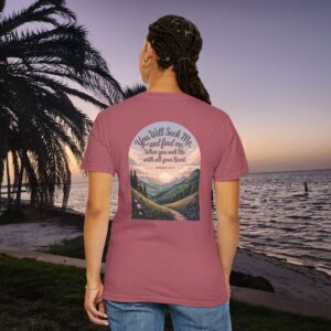 Pink Christian t-shirt with Jeremiah 29:13 scripture, mountain valley, and sunset graphic, worn by a person in a serene beach setting.