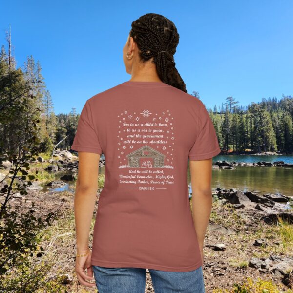 Maroon Christian t-shirt with Isaiah 9:6 scripture, stars and barn design, worn by a person standing by a lake with pine trees under a blue sky.