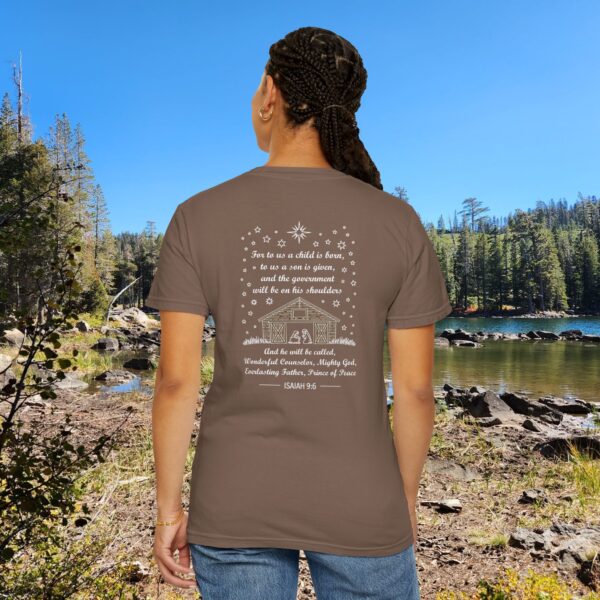 Brown Christian t-shirt with Isaiah 9:6 scripture, stars and barn design, worn by a person standing by a lake with pine trees under a blue sky.