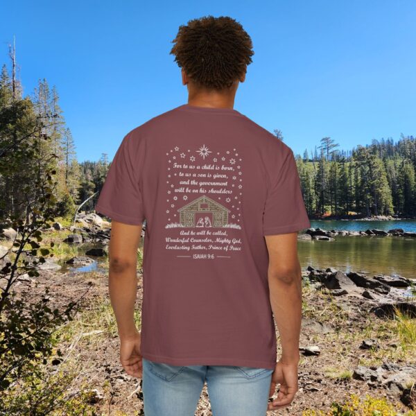 Maroon Christian t-shirt with Isaiah 9:6 scripture, stars and barn design, worn by a person standing by a lake with pine trees under a blue sky.