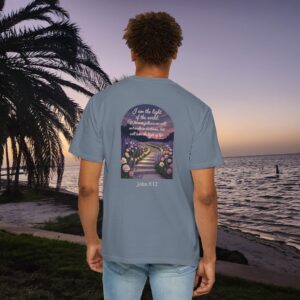 Blue Christian t-shirt with John 8:12 scripture, lit stairway path with flowers and sunset design, worn by a person standing on a sunset beach.